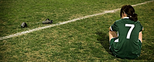 Flicka i fotbollsdräkt sitter på gräset vid fotbollsplanen.